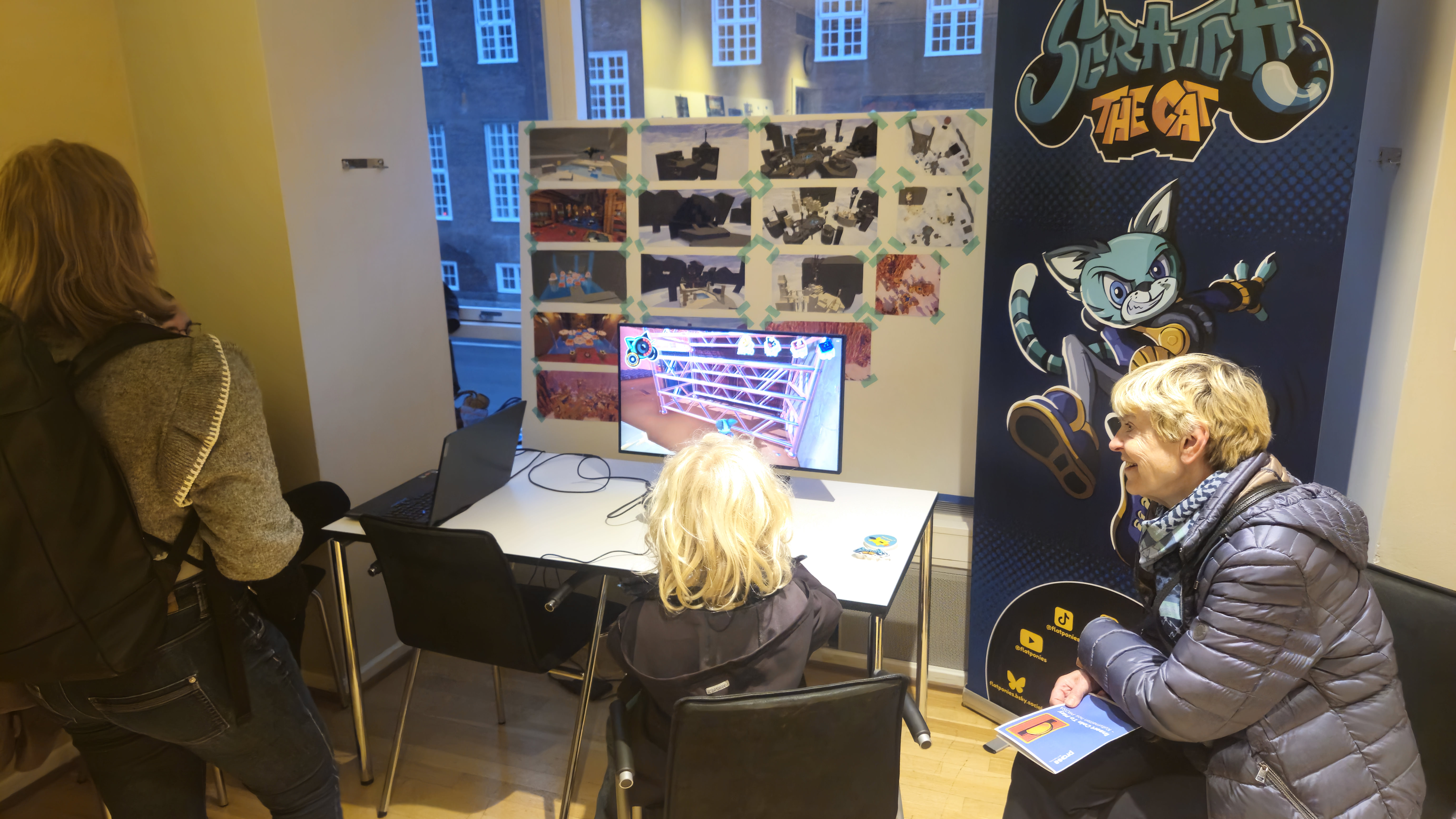 A blonde kid is sitting in a chair playing Scratch the Cat on a screen as his guardian sits to his right. Behind the screen is a big collage of all the level prototyping that FlatPonies have done and on the right is a big blue promotional poster with their Scratch character.