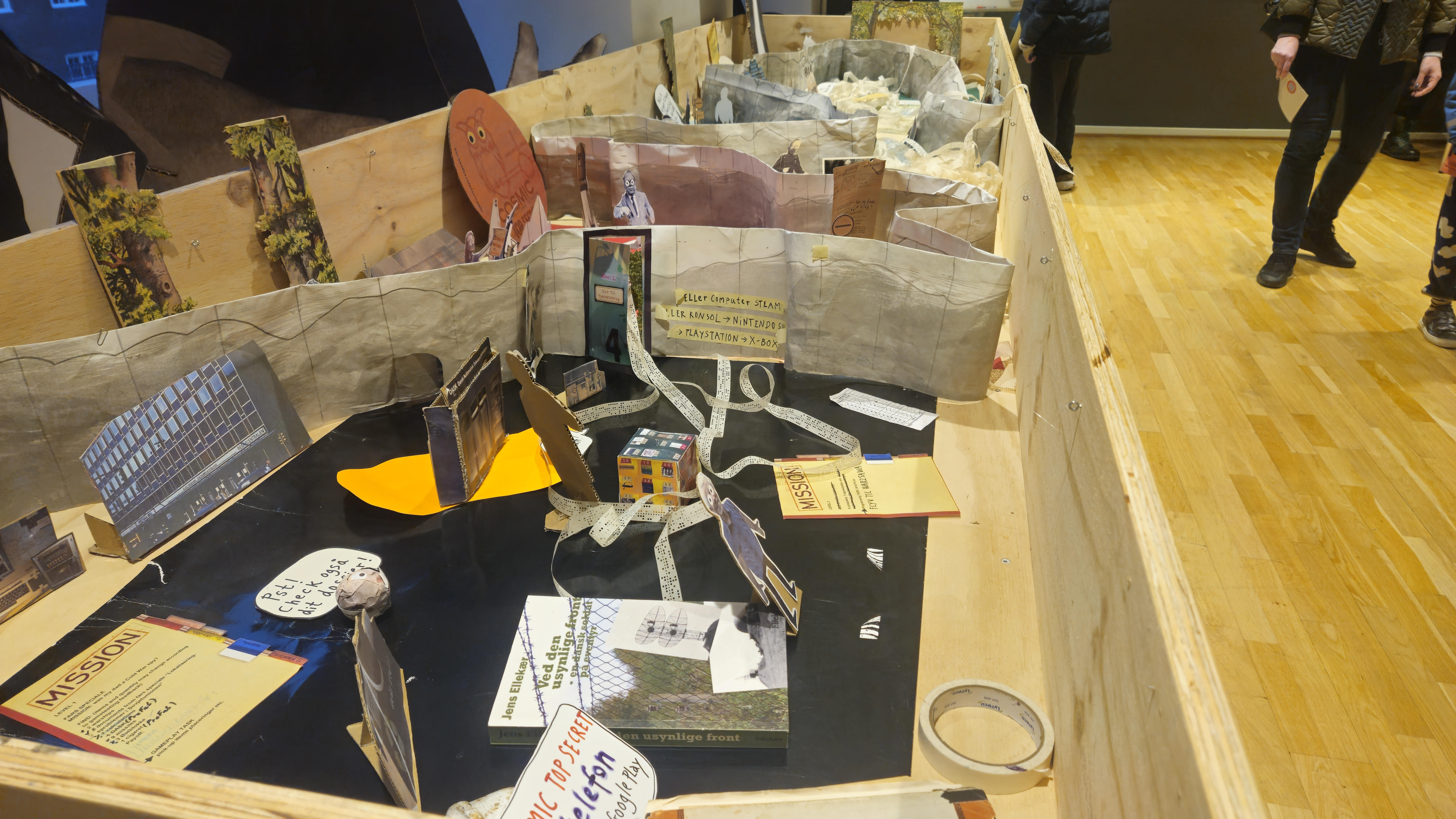 An open box display full of little figures cut from paper, black surfaces for flooring, long strips of paper with holes in produced by an old Danish government computer called DASK as well as paper walls with writings on them.