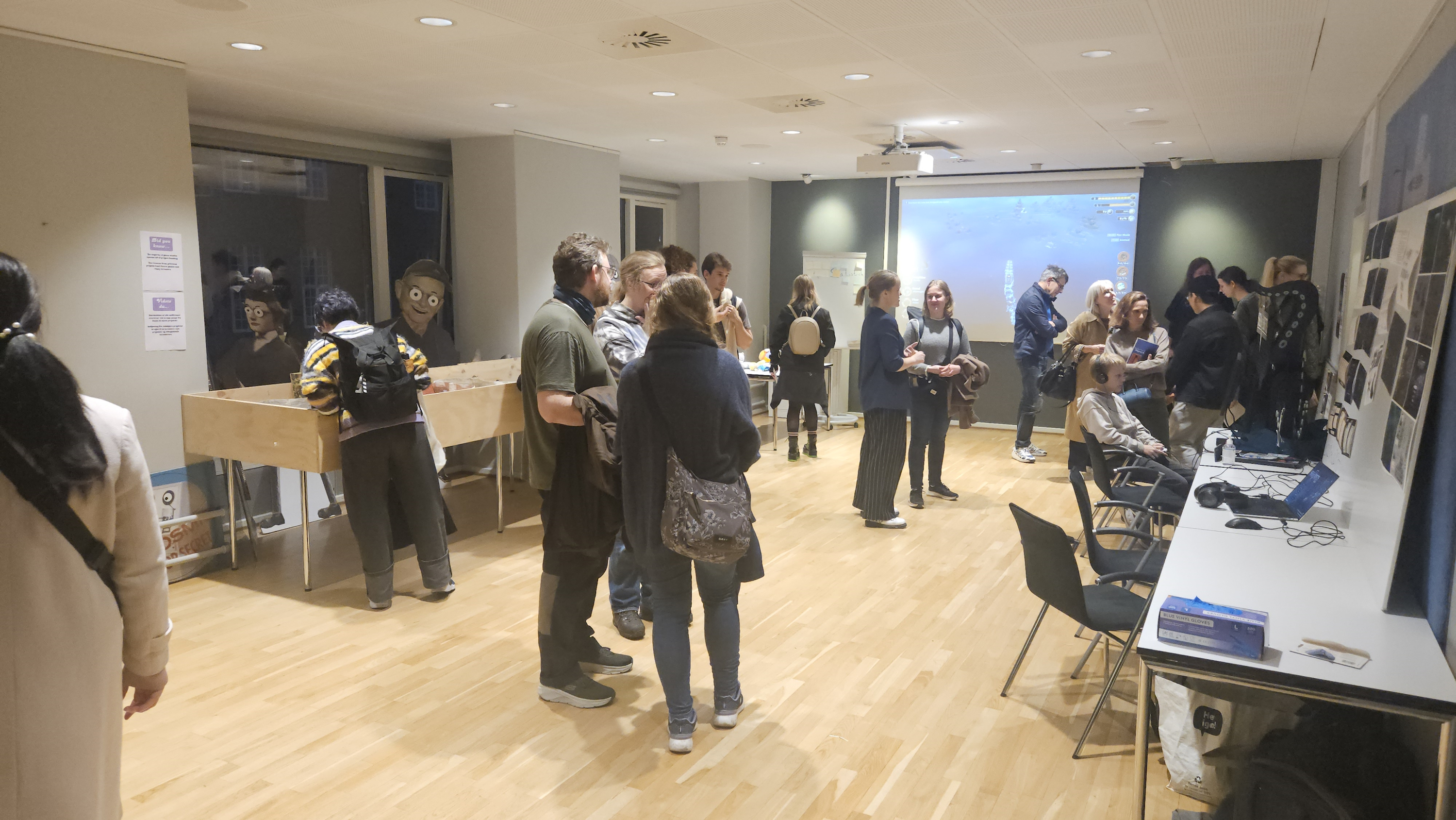 A room full of people in PROSA's canteen checking out the various exhibits, playing games and asking questions or talking to organisers and developers. In the left part of the background is an exhibit by the company Those Eyes and in the right side of the background is a big projector screen running various trailers from all the game companies we showcased material from today.