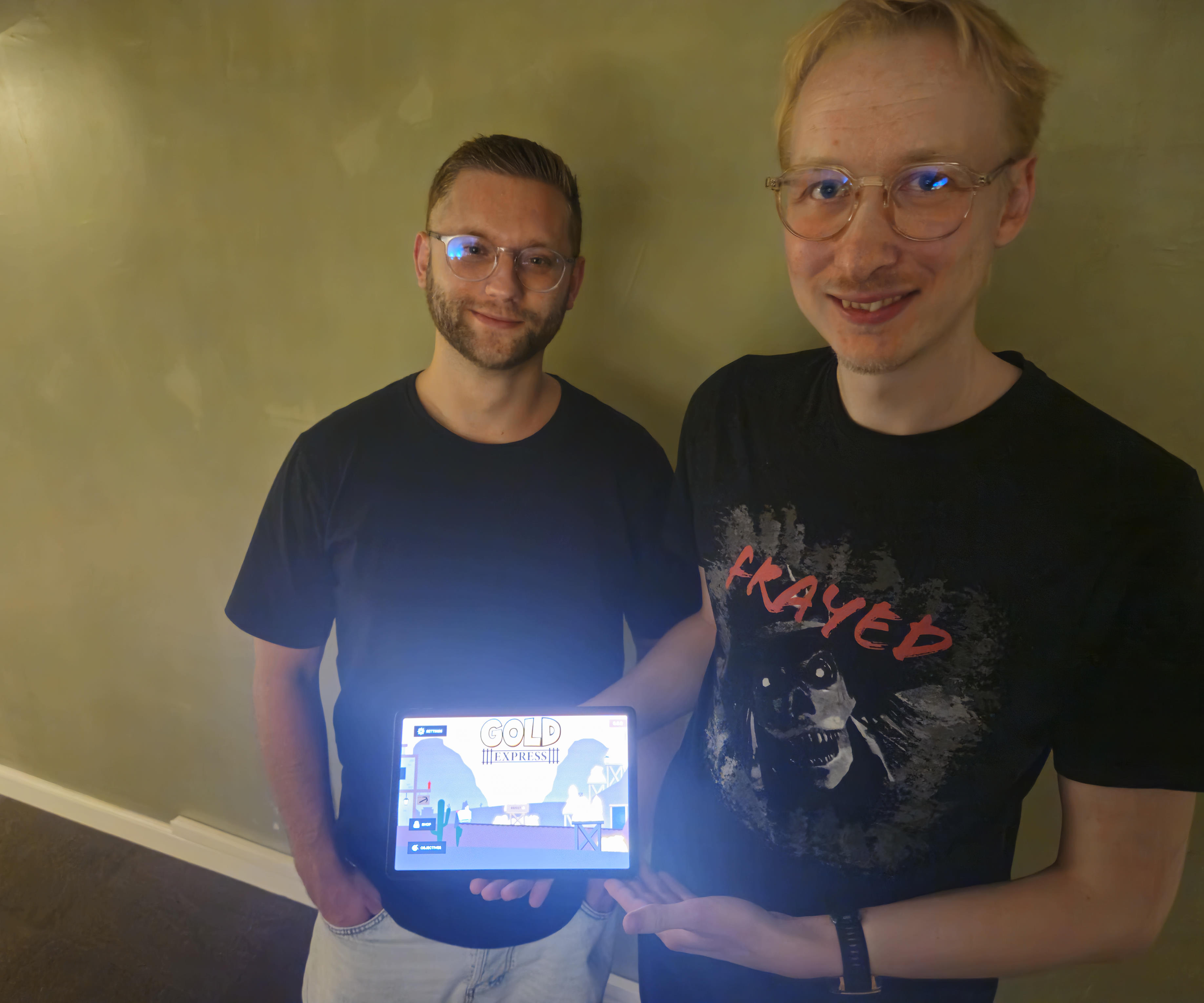 Aleksander Willersrud (Left) and Marcus Due Jensen (Right) standing against a wall with their games title screen visible on a table that Marcus is holding.