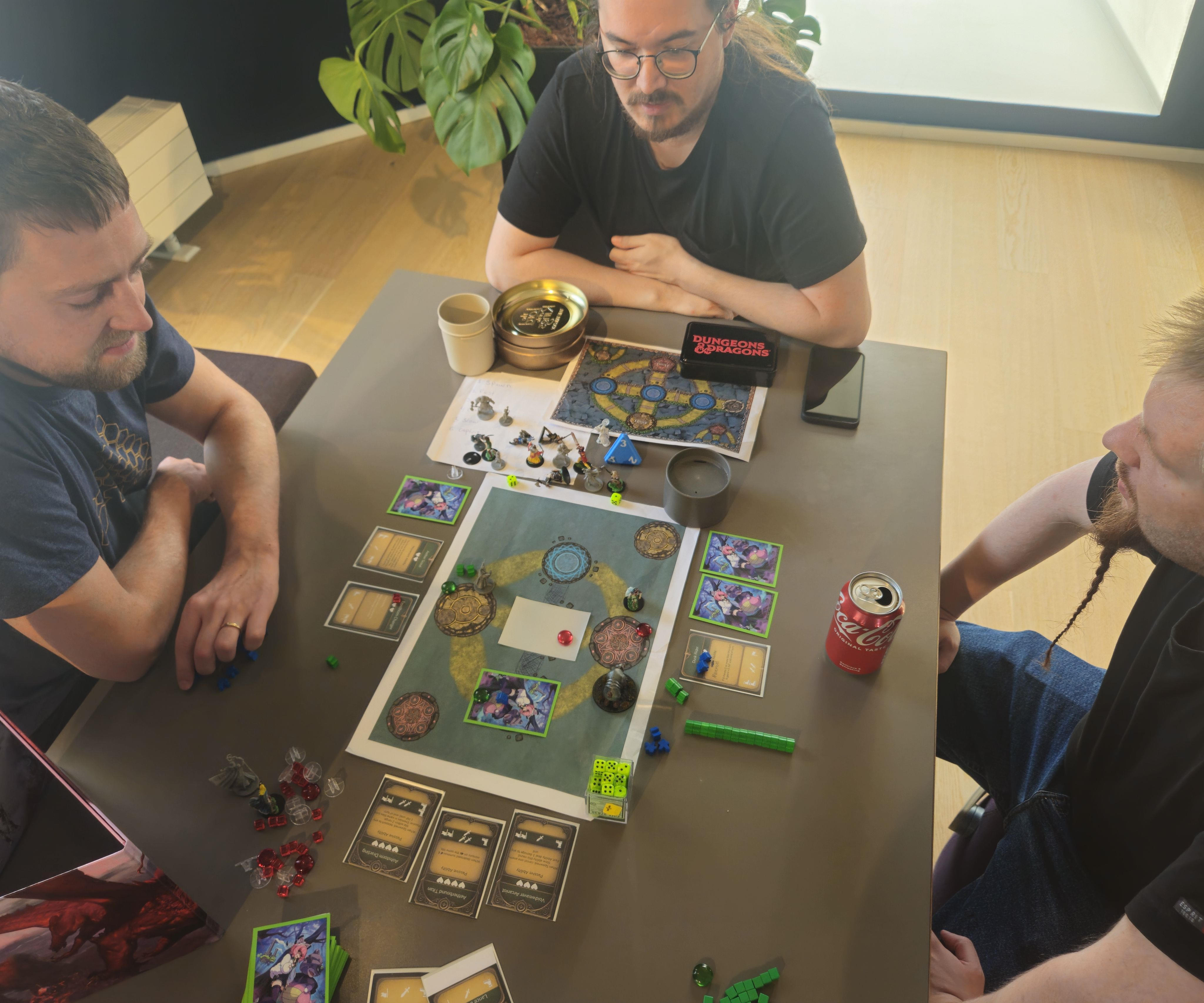 On either side of a table sits two players playing against each other while the games creator sits off to one side looking over the game and controlling certain aspects of it like a game master.