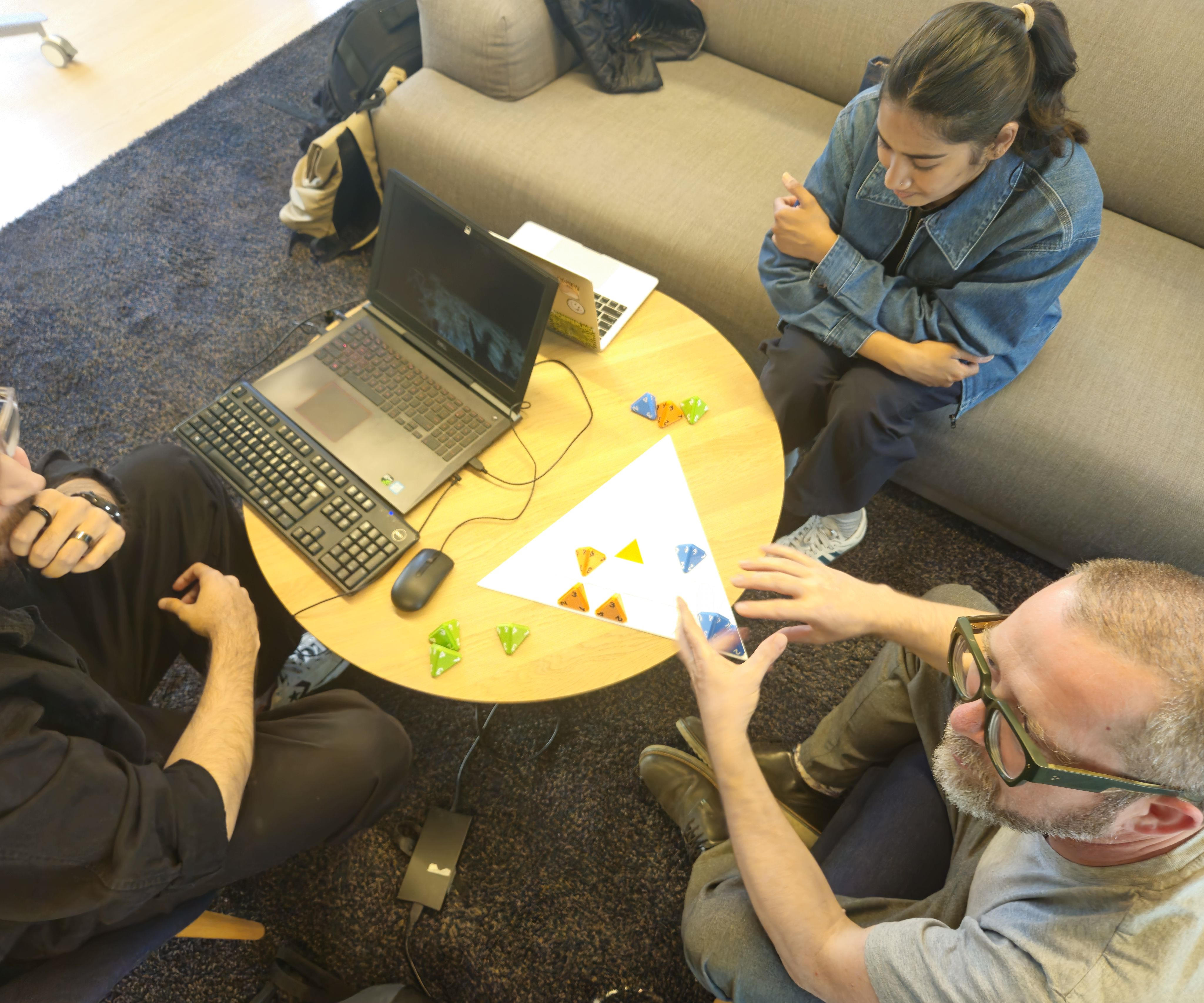 Three players sitting around a triangle shaped board with triangle shaped divets in it to hold four sided dice. One player has already been beaten out of the game and the creator is explaining a concept to the remaining player.
