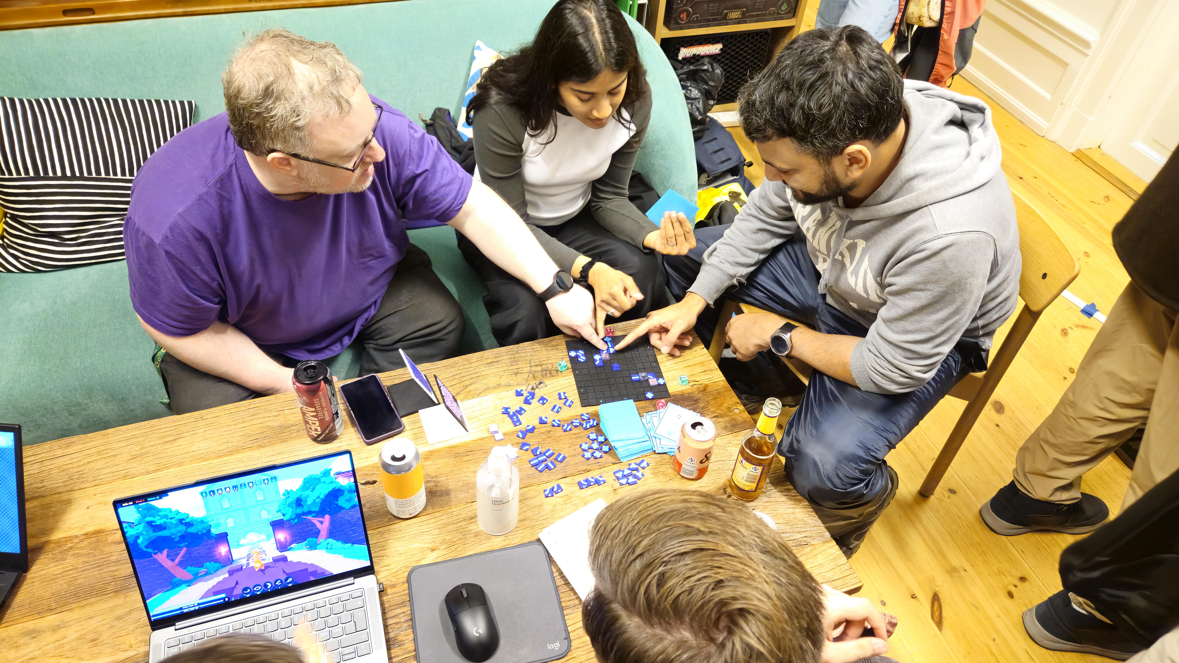 Kim (Left) sitting in his purple shirt explaining a rule to two other players, a woman sitting in the middle and a man sitting out to the right.