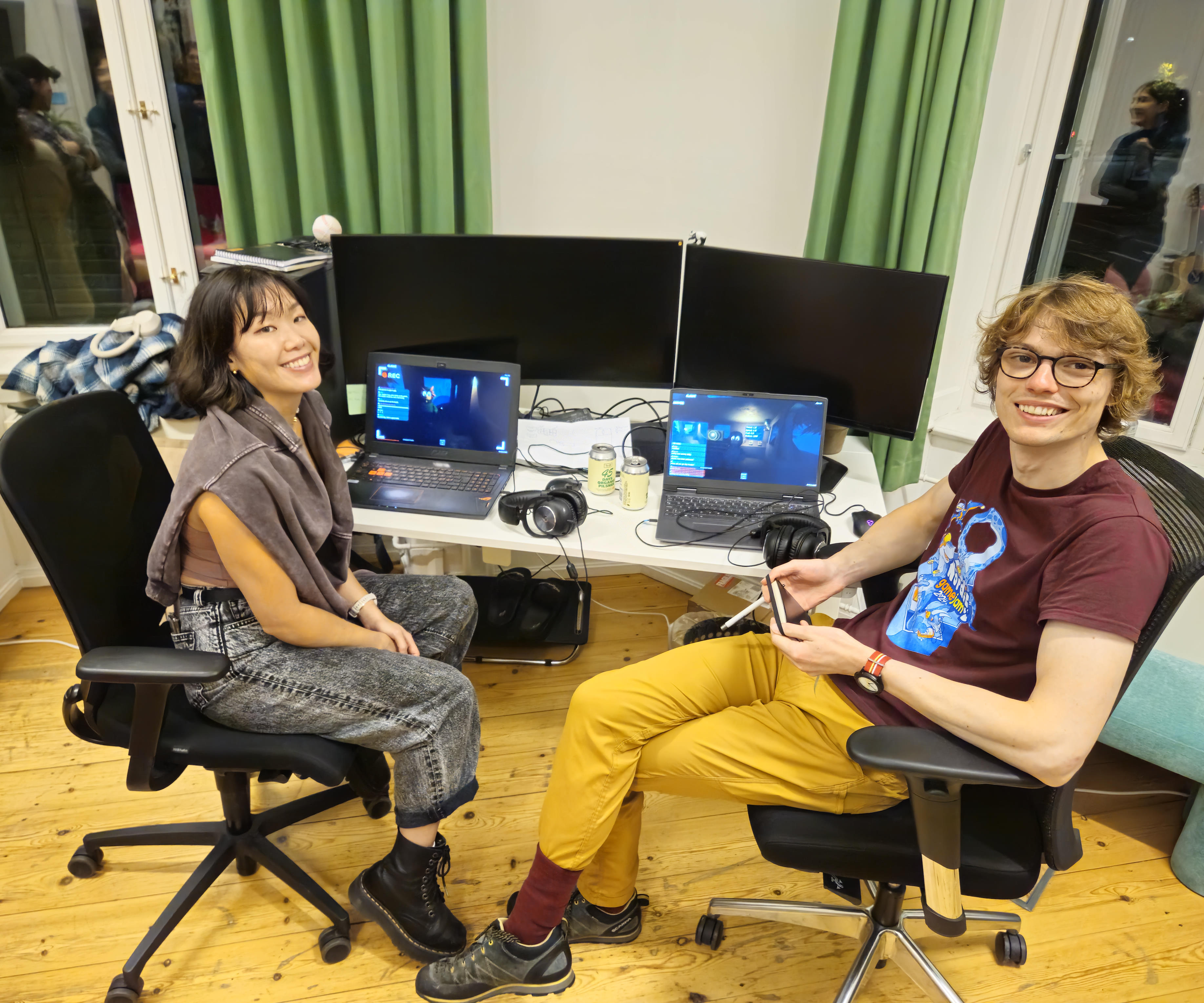 Game Designers Teresina Ho (Left) and Max Wrighton (Right) sitting in front of their laptops smiling at the camera with the game running in the background.